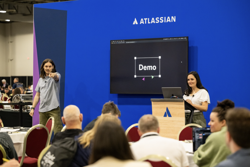 Two Atlassian employees present a demo to an audience of peers with a large screen between them. 